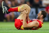 A Gold Coast Suns' AFL player grimaces in pain as he lies on the ground holding his left leg.
