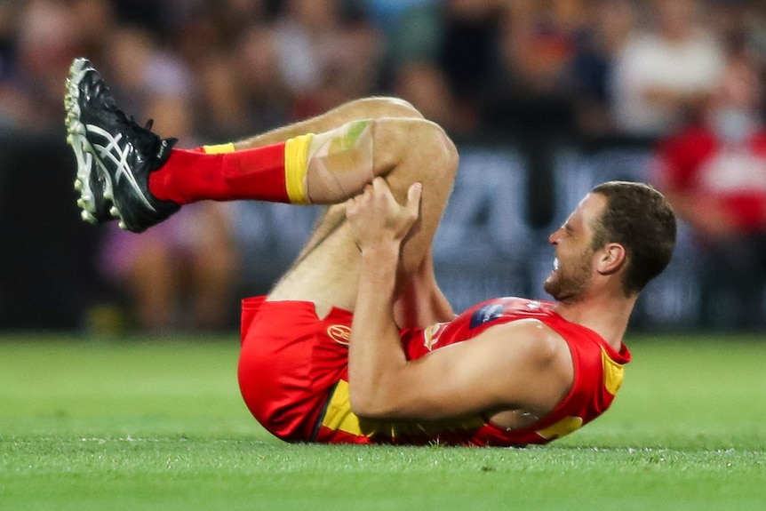 A Gold Coast Suns' AFL player grimaces in pain as he lies on the ground holding his left leg.