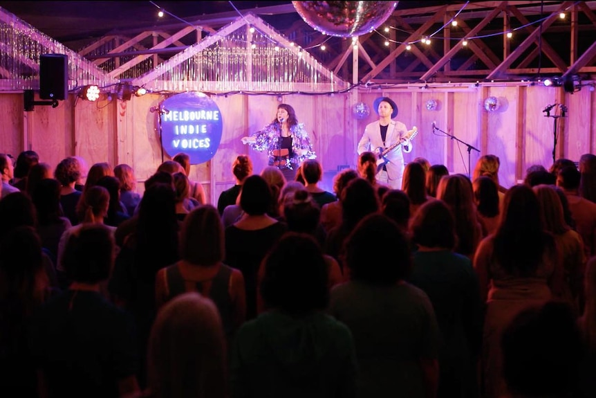 a crowd of people facing a stage at the back where a woman directs the crowd and a man plays guitar.