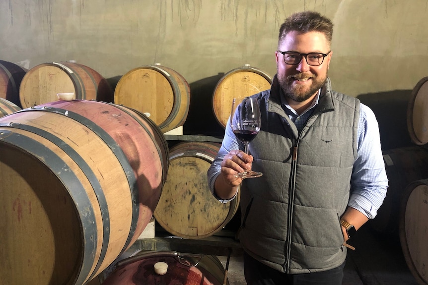 A man wearing a blue shirt and grey vest holding a glass of wine next to a wine stained wine barrel