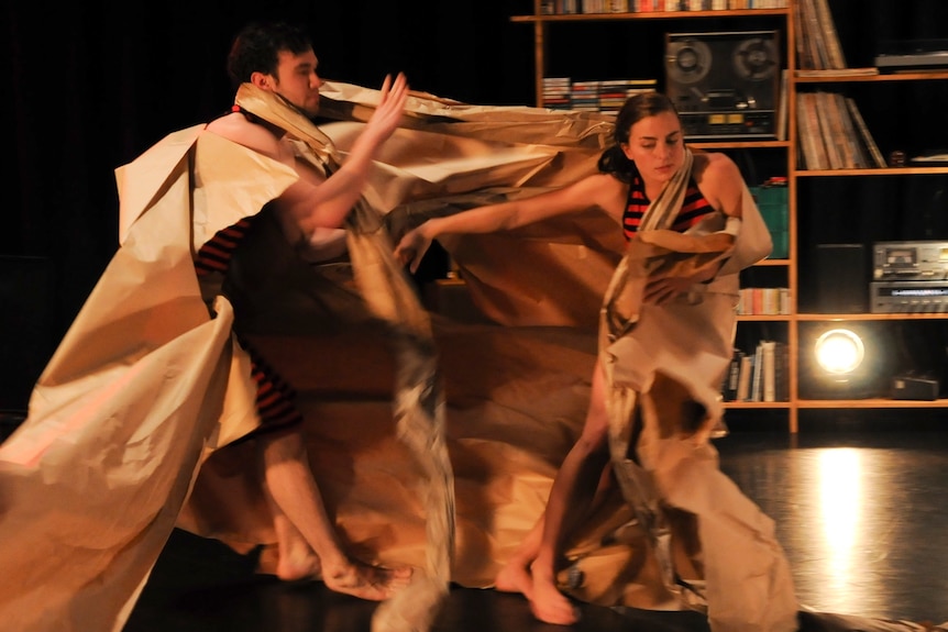 A white man and woman dance on a stage, entwined in brown paper wrapping and with a bookshelf in the background.