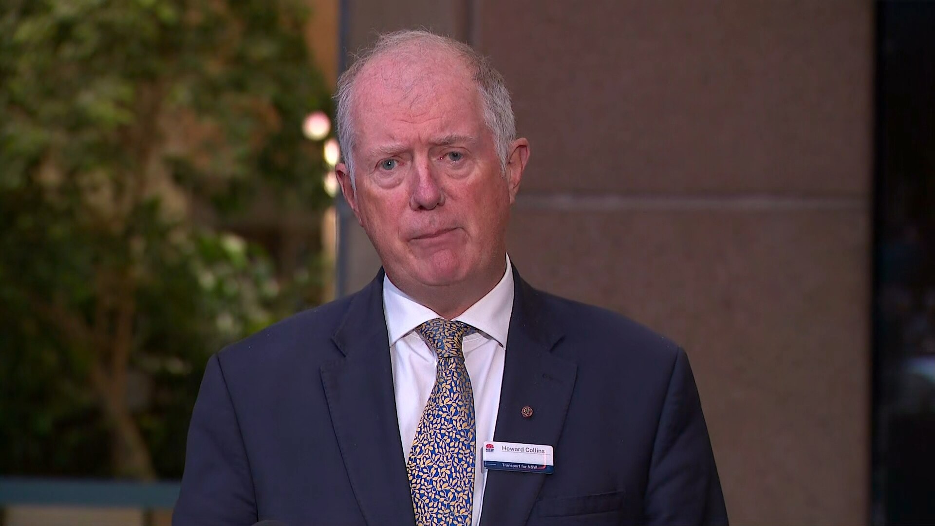 Un hombre mayor con un traje de pie frente a una pared de ladrillo, hablando.