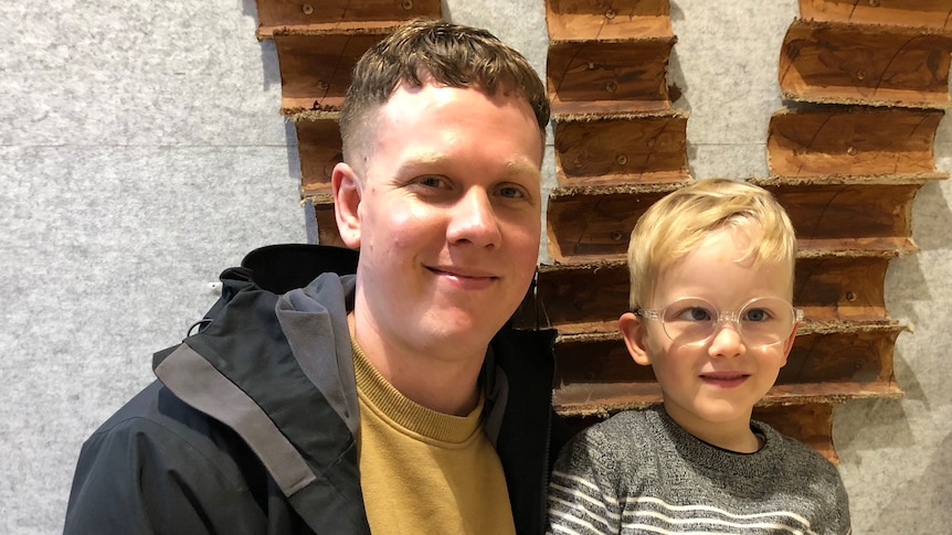 A man, wears black jacket, mustard jumper,  blond boy, wears glasses, smile, stand in front of wooden installation on wall.