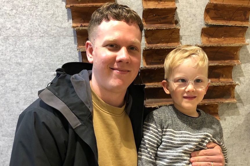 A man, wears black jacket, mustard jumper,  blond boy, wears glasses, smile, stand in front of wooden installation on wall.