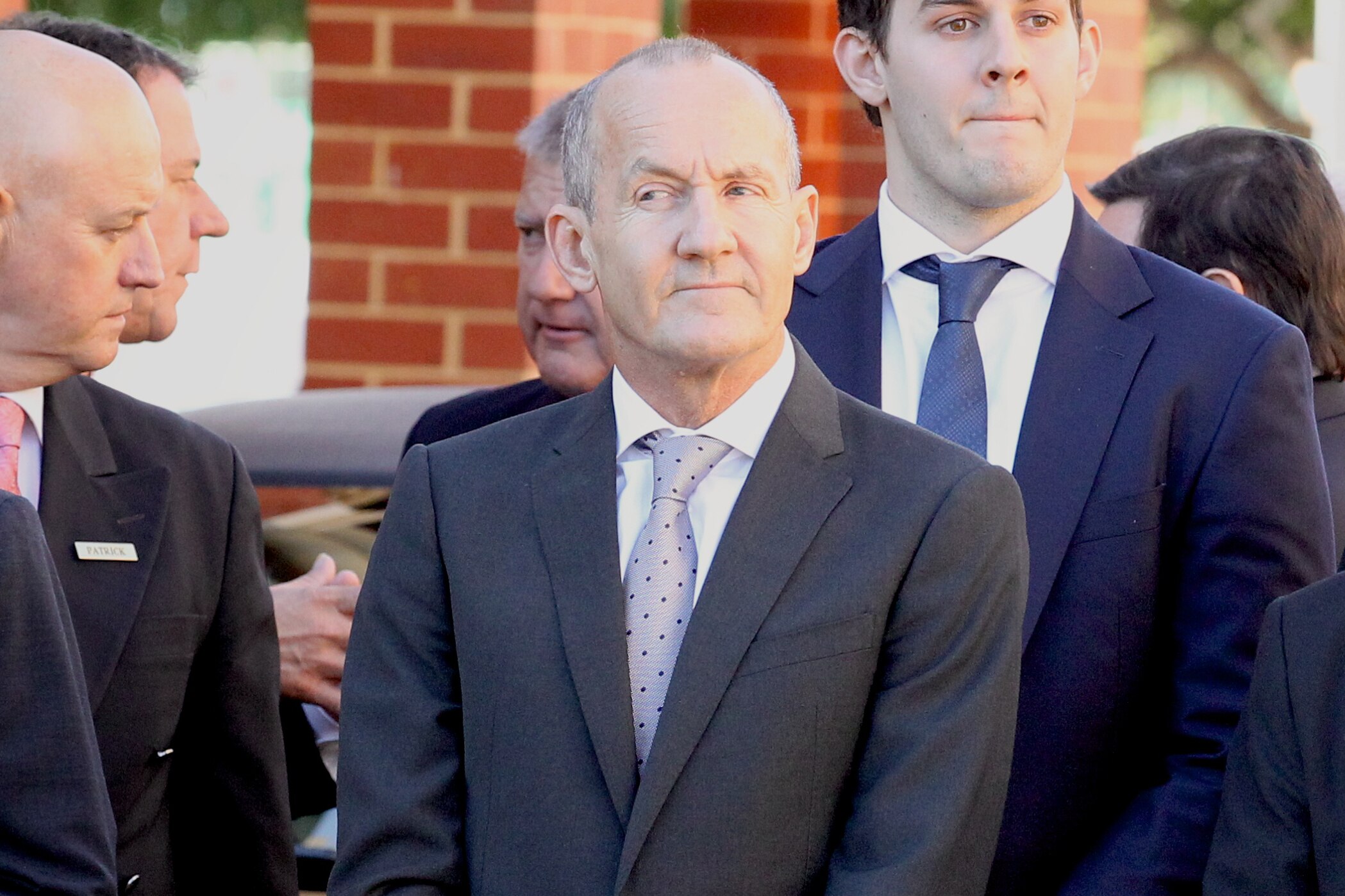 A mid-shot of John Bond dressed in a suit and tie glancing sideways in front of a group of men at a funeral.