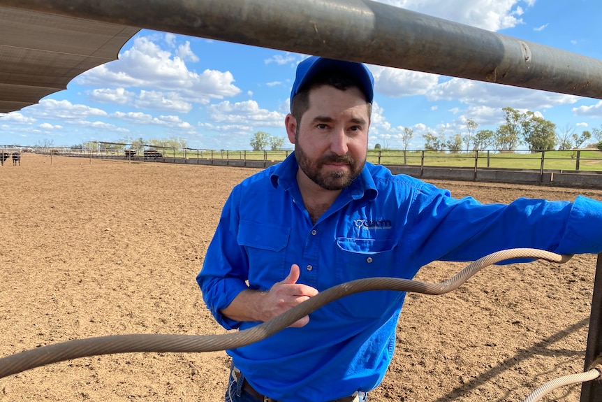 Farmer wearing a blue shirt and cap, standing behind a fence.