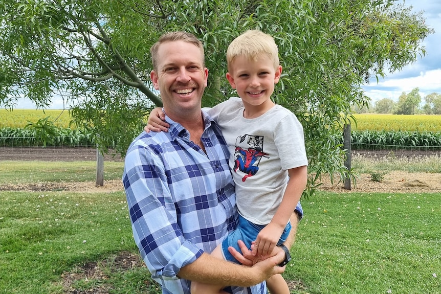A man wearing a checked shirt holds his son in his arms in a rural setting.