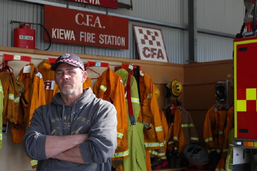 A man wearing a CFA cap standing in front of a rack of fire fighting jackets, equipment and Kiewa fire station sign.