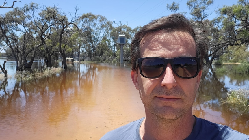 A man with short, dark hair stands on the bank of a swelling river.