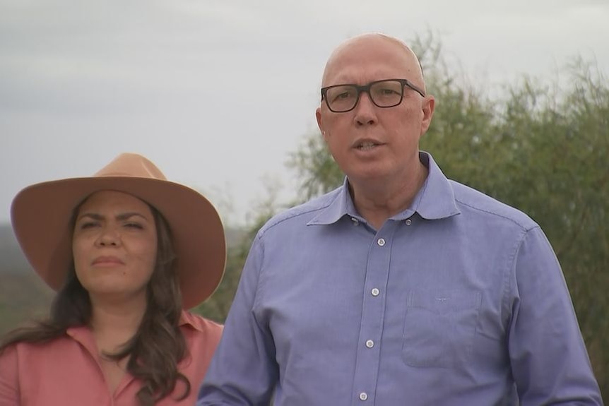 Jacinta Price wearing a hat and squitnting standing outside next to Peter Dutton who's wearing a blue shirt and black glasses