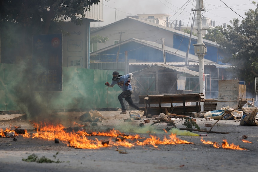 A man wearing a Superman T-shirt runs past a road barricade and burning debris.