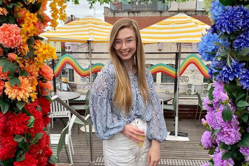 A woman, smiling with glasses, posing for a photo amongst a floral display. 