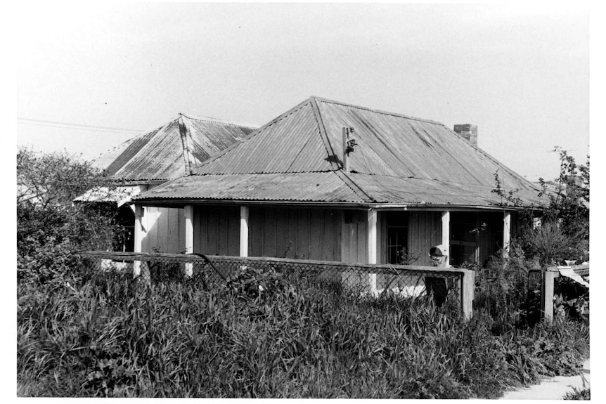 A black and white image of a cottage with long grass or shrubs surrounding it.