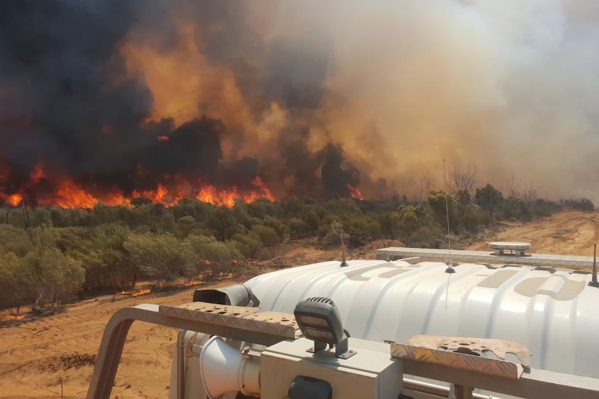 A fire vehicle sits in the foreground as a fire burns through bushland in the background.