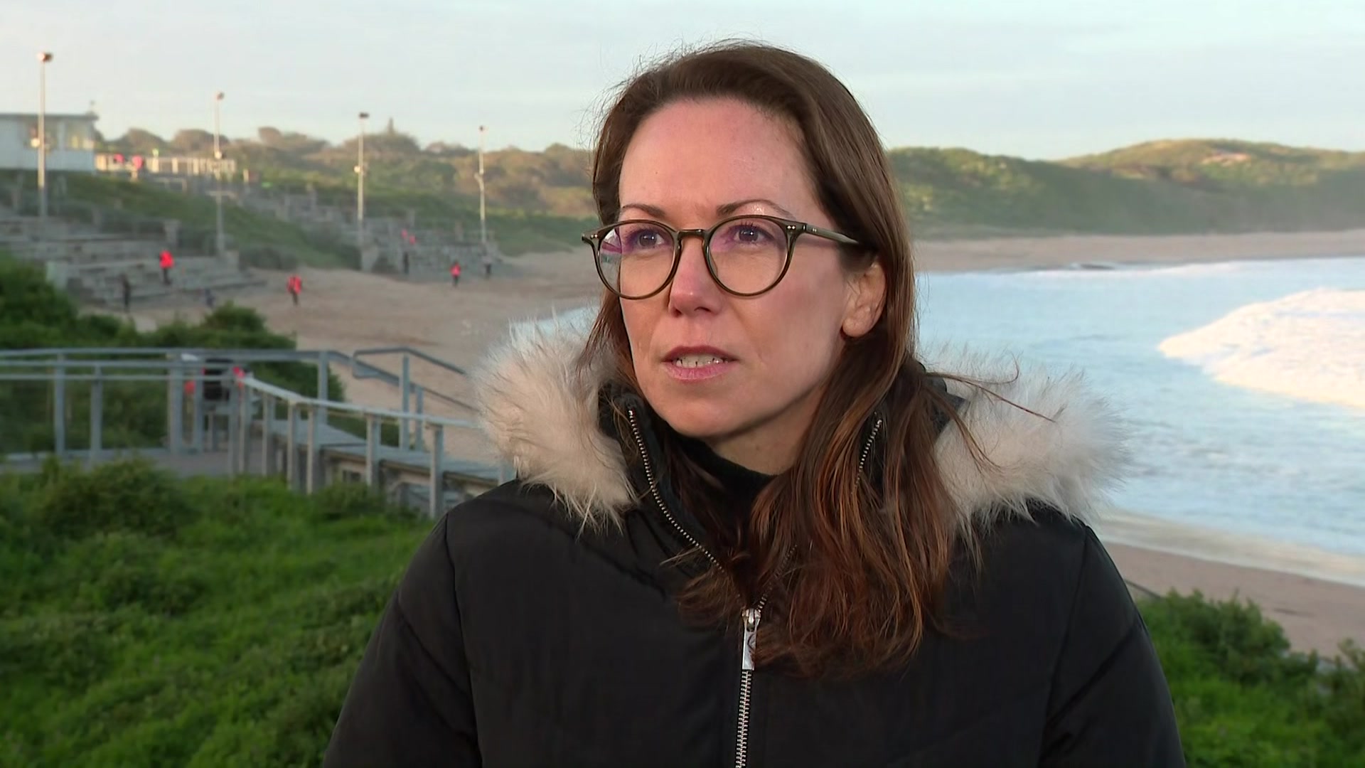 A dark haired woman wearing glasses and a black jacket with a fur-lined hood stands near a beach where waves are rolling in.
