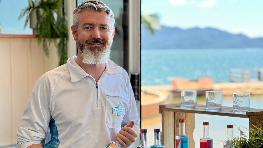 A man smiles in front of a window with an island and ocean view holding a bottle of gin.