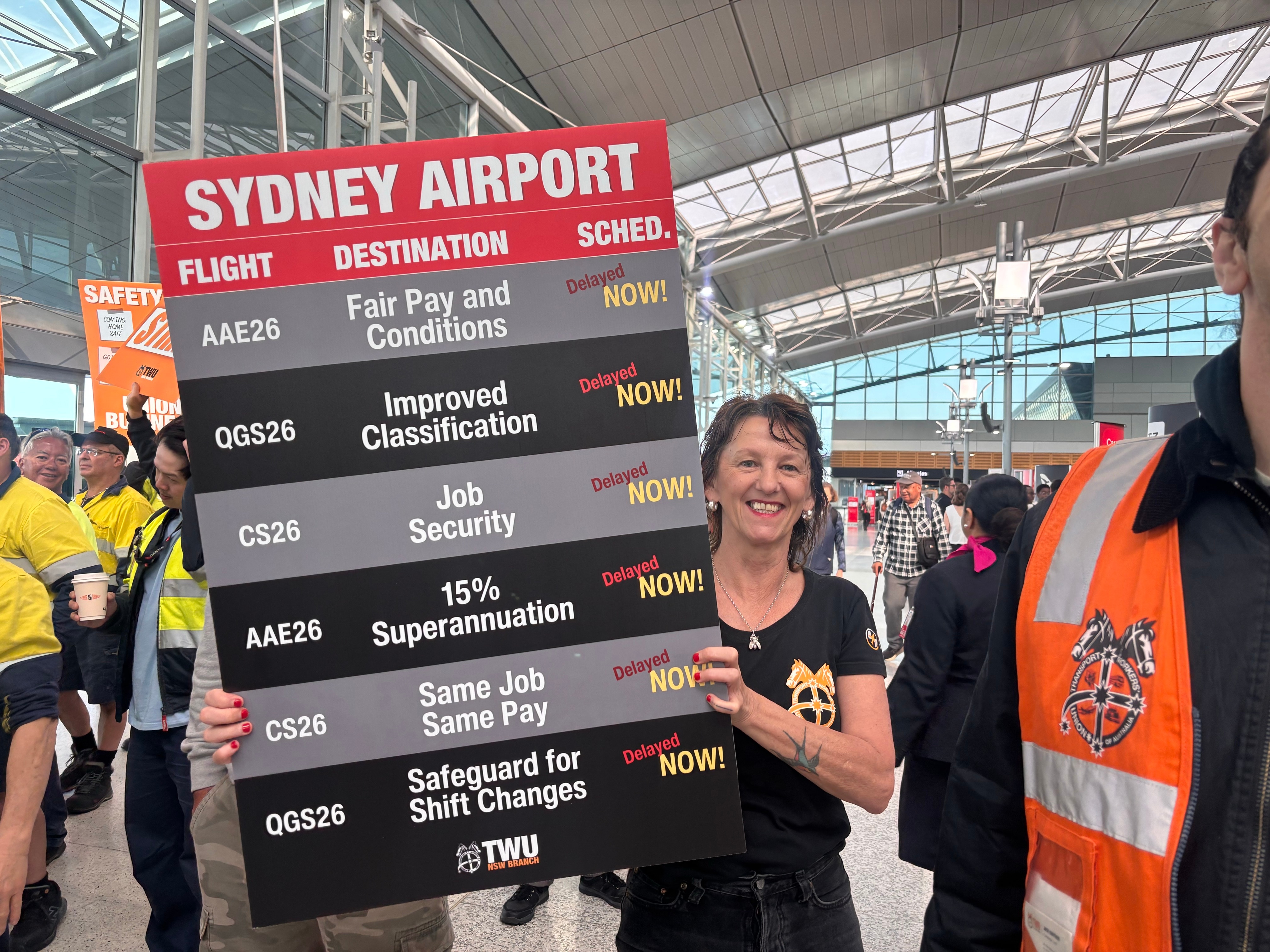 A woman with short hair smiles holding a large sign demanding better working conditions for airport ground crew.