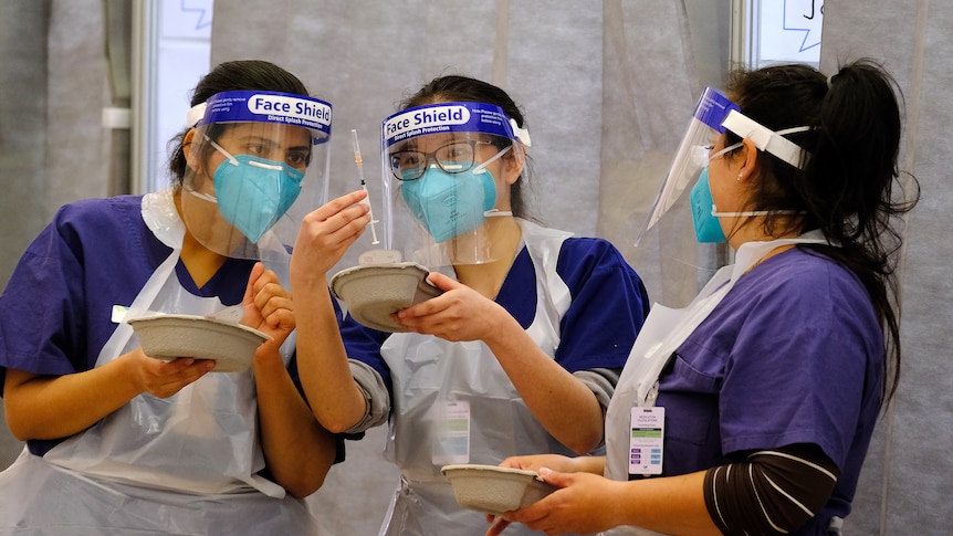 Three nurses in PPE inspect a needle.