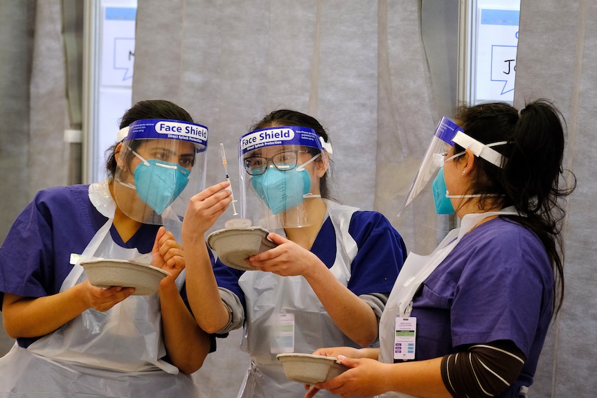 Three nurses in PPE inspect a needle.