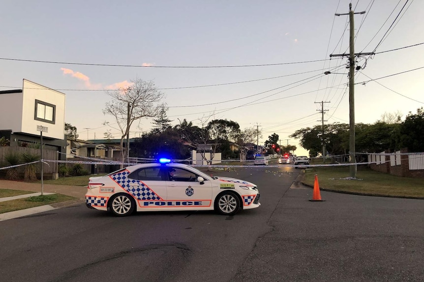 A police car with lights on blocks a suburban Brisbane street at sunrise, police tape also closes the road.