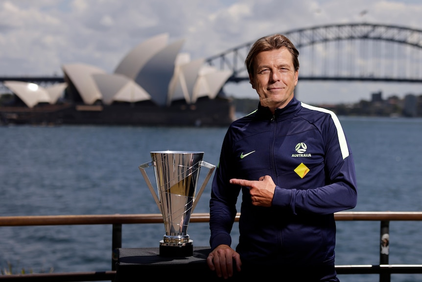 A man wearing a navy blue sports jacket points to a trophy in front of some tourist attractions in Australia
