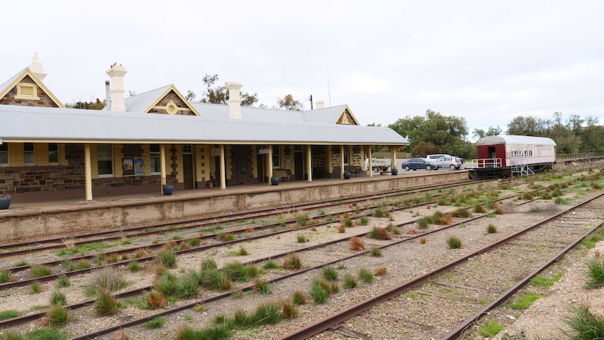 A brick-built railway station sits beside a disused track covered in weeds and the dining carriage can be seen.