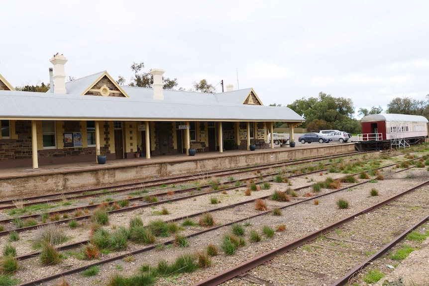 A brick-built railway station sits beside a disused track covered in weeds and the dining carriage can be seen.