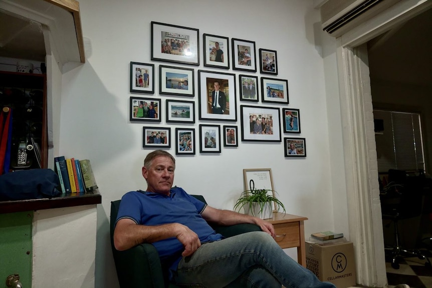 A father sits on a chair in front of family photos.