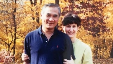 A smiling woman hugs a smiling man as they stand near trees and shrubs in yellow and red autumn colours.
