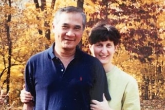 A smiling woman hugs a smiling man as they stand near trees and shrubs in yellow and red autumn colours.