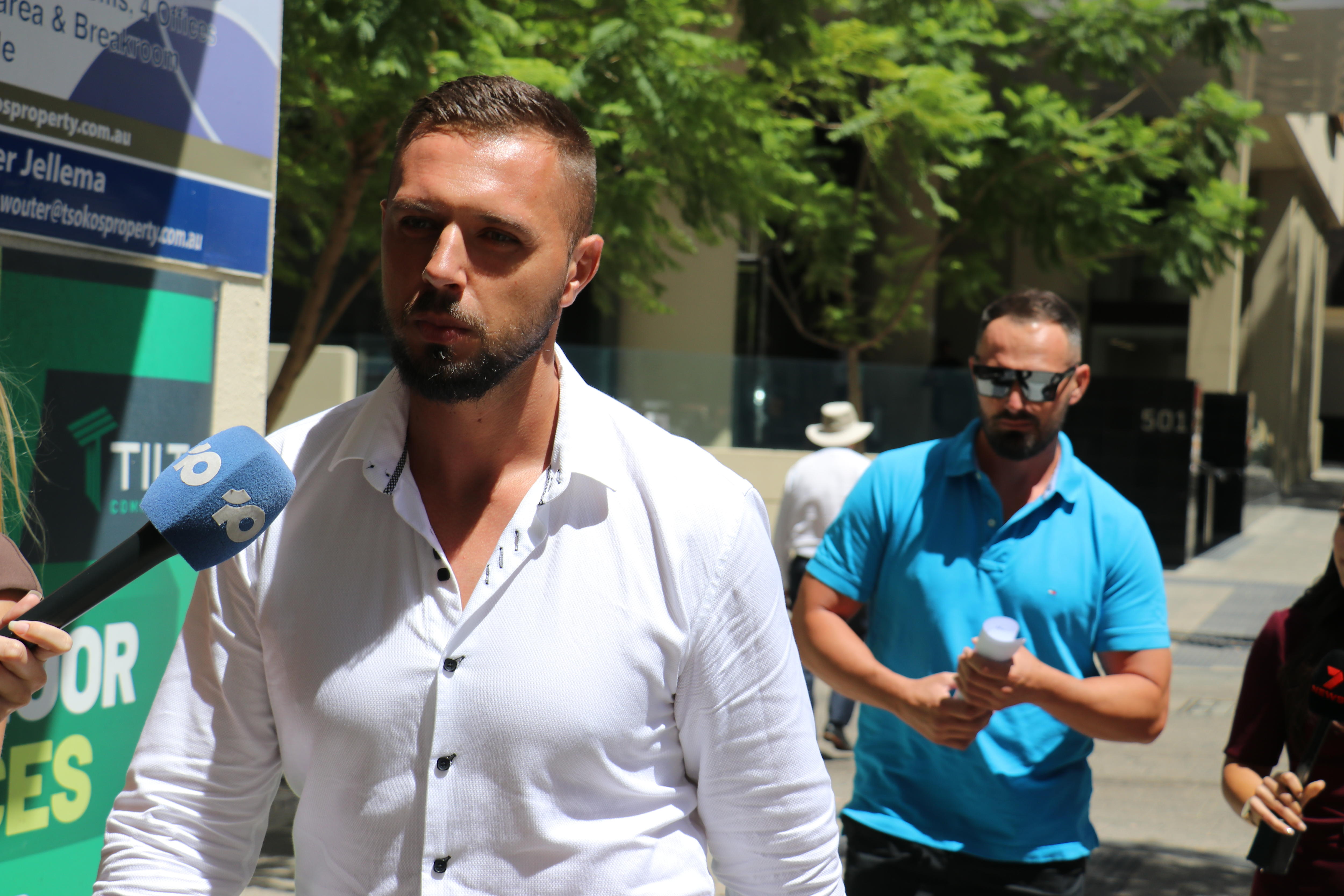 Un plano medio de Nezir y Enver Ibrahimovic saliendo de la sala del tribunal, con un micrófono de reportero apuntando a Nezir a la izquierda. 