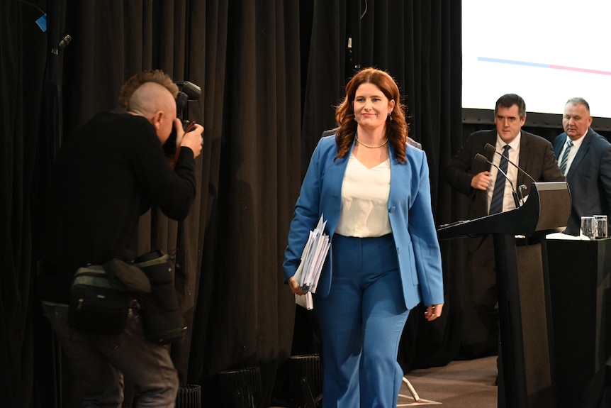 A woman in a blue blazer and suit walks towards a photographer, in front of black curtains