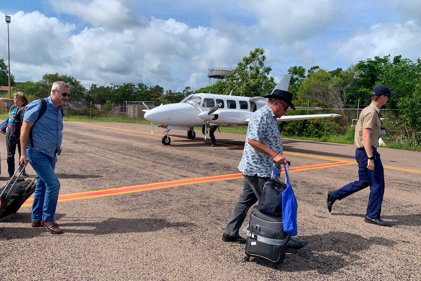 Mick Dodson walks across the tarmac at a small remote airport, wearing an Akubra had and wheeling a small piece of luggage.