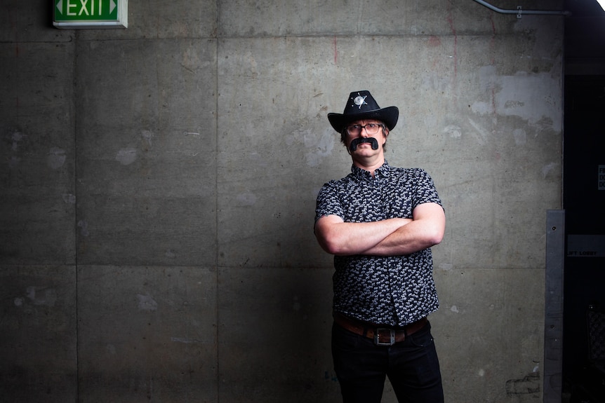 A man in a fake moustache and cowboy hat stands in front of a grey wall.