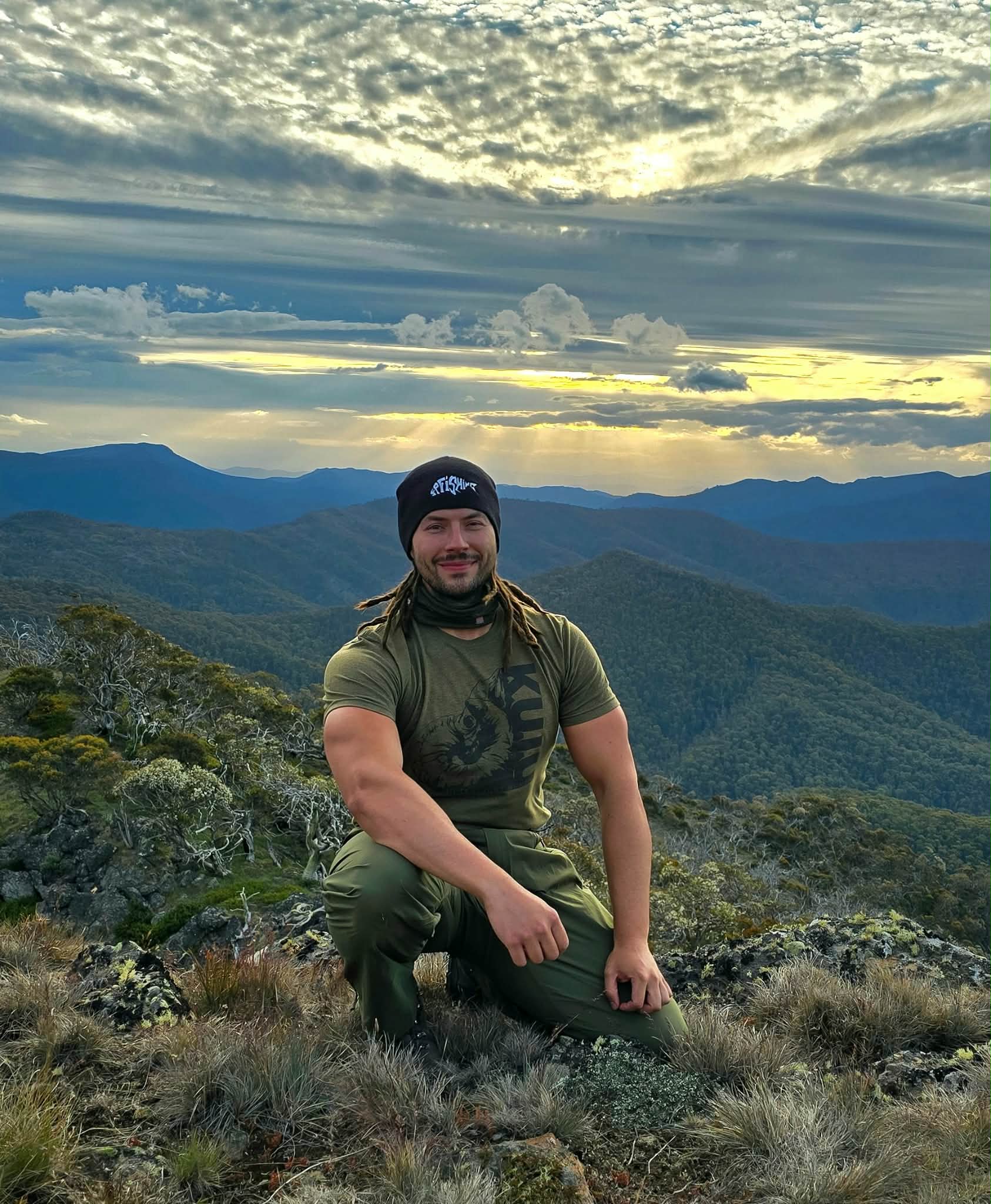Un hombre vestido con una camiseta verde, pantalones verdes y una gorra negra en la cabeza se arrodilla en una montaña