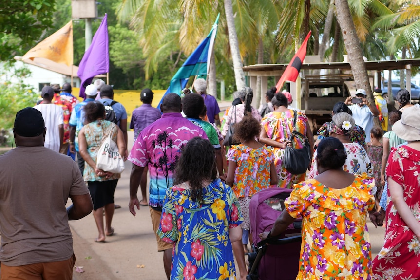 A group of people walk in colourful floral dresses and shirts, some with flags