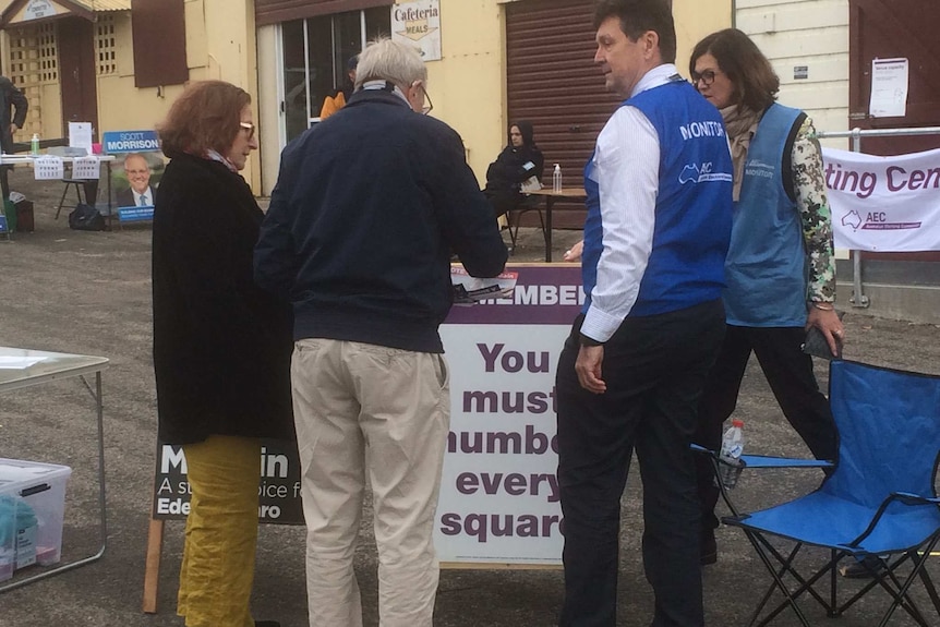 A man in an official's vest speaks with a man and a woman at an outdoor polling station.