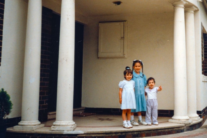 An older photograph of three little girls standing on the steps outside a home, flanked by white pillars