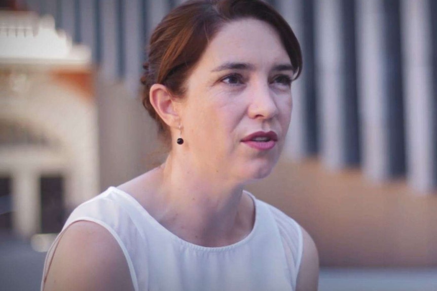 Erica Barrenger wearing a white sleeveless top, speaking in a courtyard in the city.