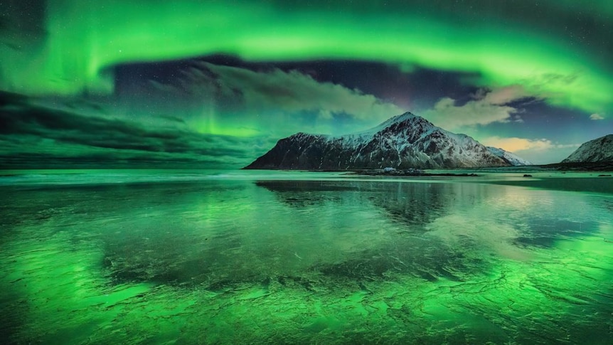 A green aurora in the sky and is also reflected off the beach with a snowy mountain in the background