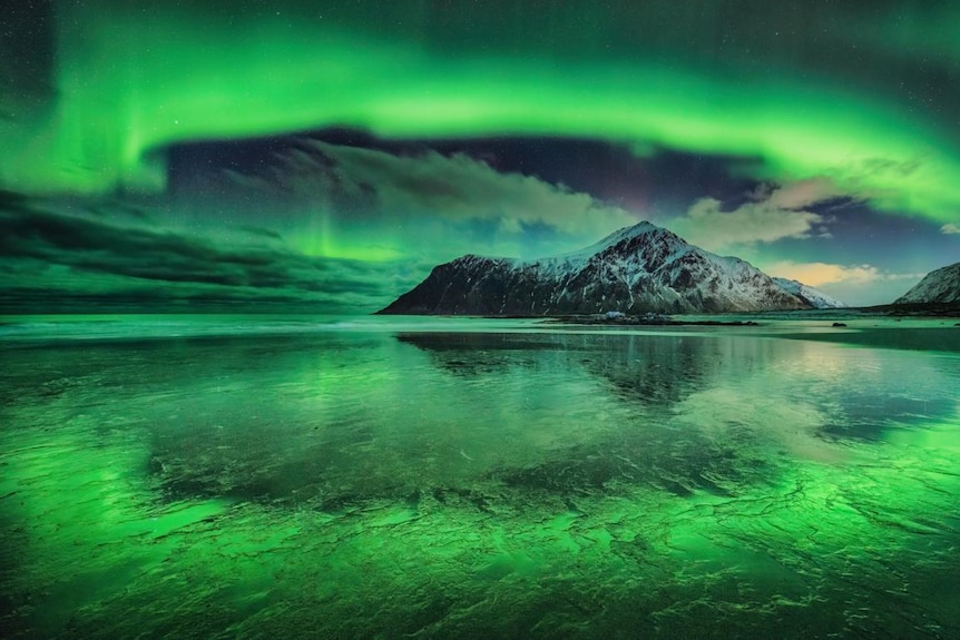 A green aurora in the sky and is also reflected off the beach with a snowy mountain in the background