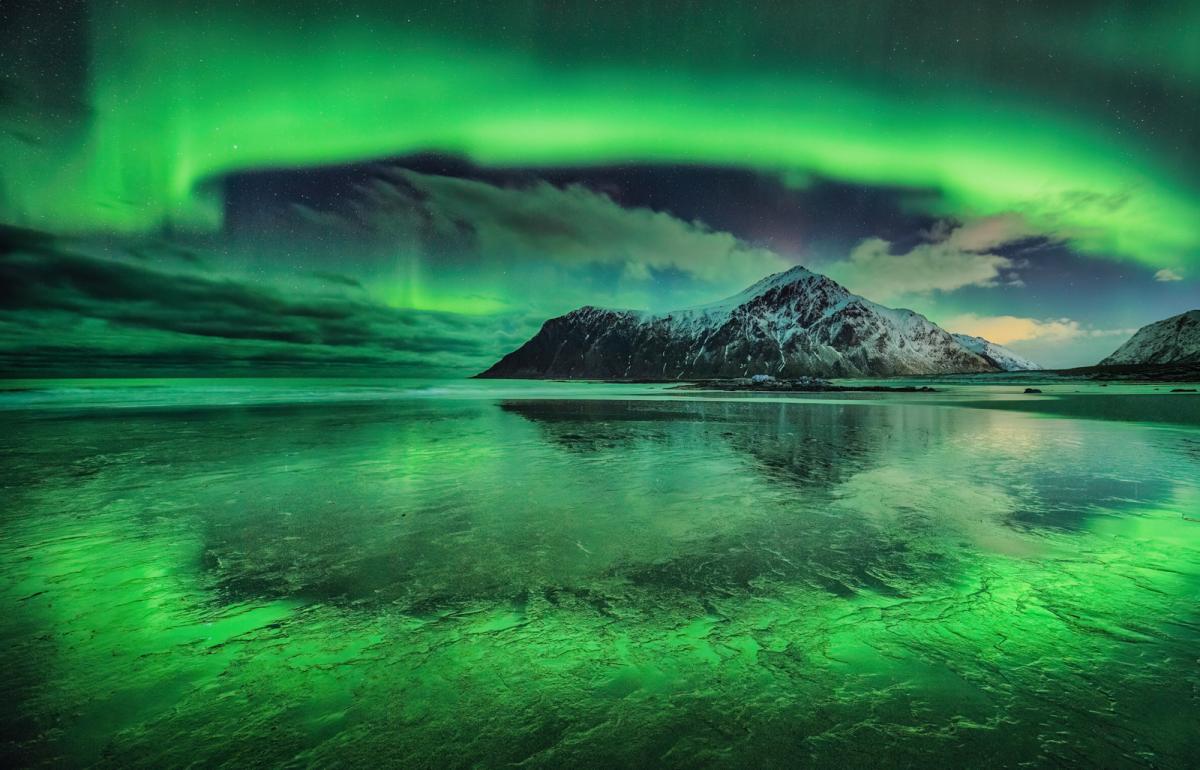 A green aurora in the sky and is also reflected off the beach with a snowy mountain in the background