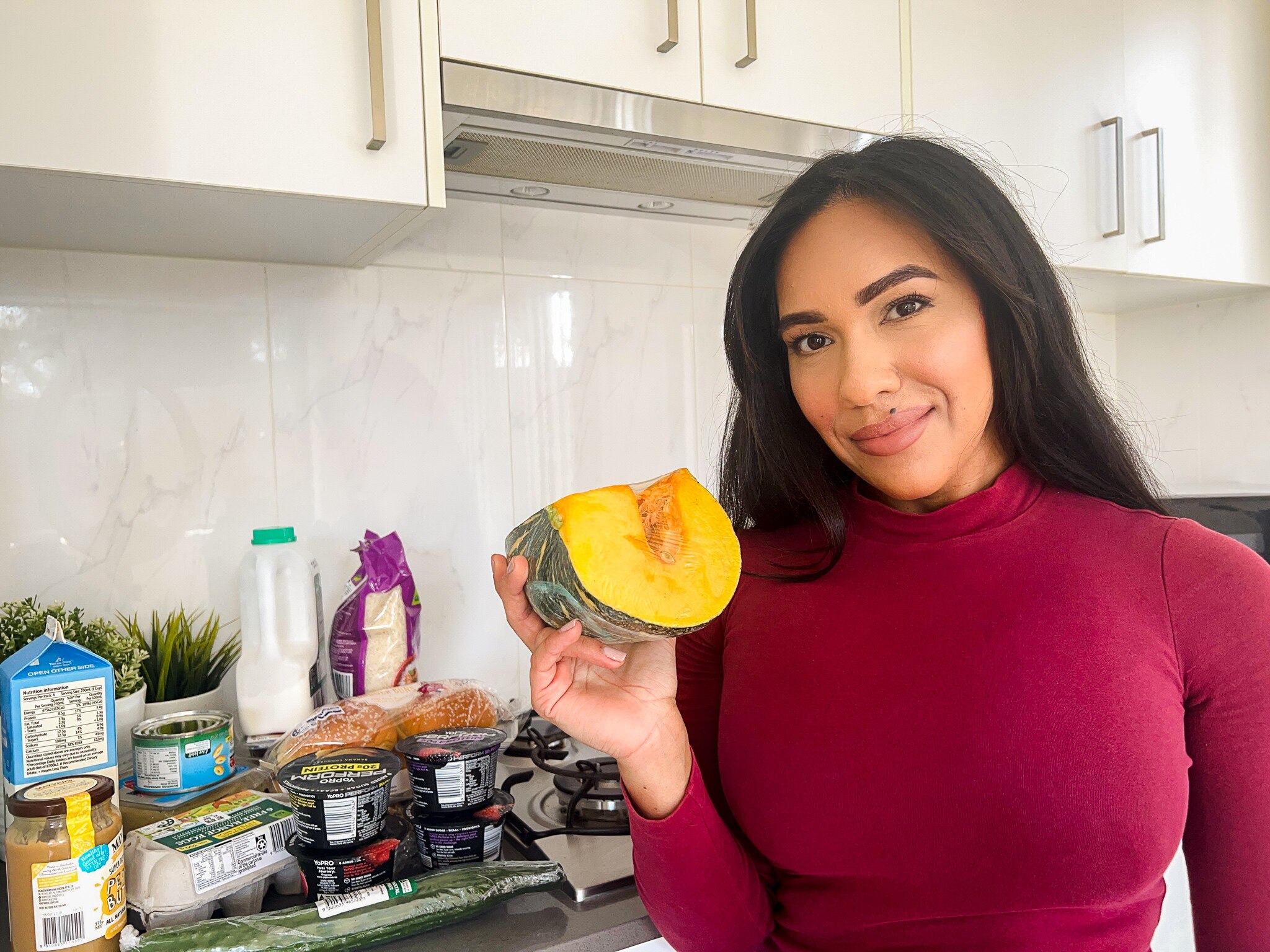 Tahnee holds a pumpkin in her right hand standing next to her grocery shopping in the kitchen.