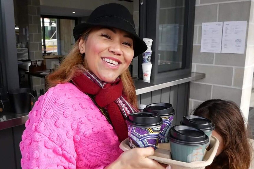 A woman in a bright pink jumper and black hat smiles at the camera carrying four takeaway coffees in a cardboard holder.