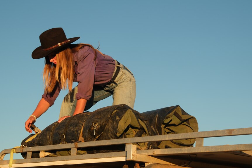 a young woman in jeans and a button up purple shirt and black cowgirl hat stands on top of a ute tying down a swag