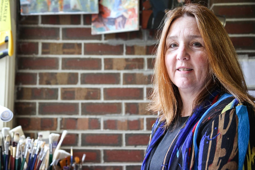 A smiling woman stand in room with paintbrushes and artworks.