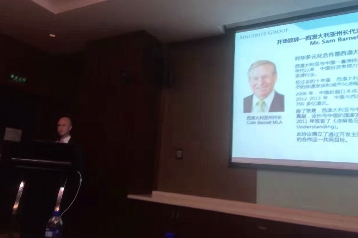 Sam Barnett at a lectern with a picture of Colin Barnett on a screen.