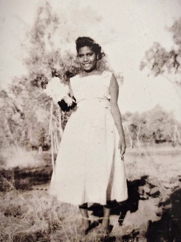 A young Aboriginal woman stands in a bush setting in a white dress holding a bouquet of flowers