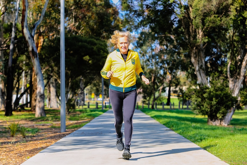 A woman in a yellow track suit top jogging in a park.