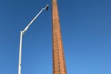 Looking up at tall brick chimney from the ground as a white cherrypicker expands to scale it against a blue sky.